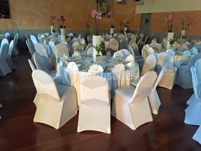 Salle de réception avec des tables rondes dressées, nappes blanches et chaises couvertes, décorées d'orchidées.
