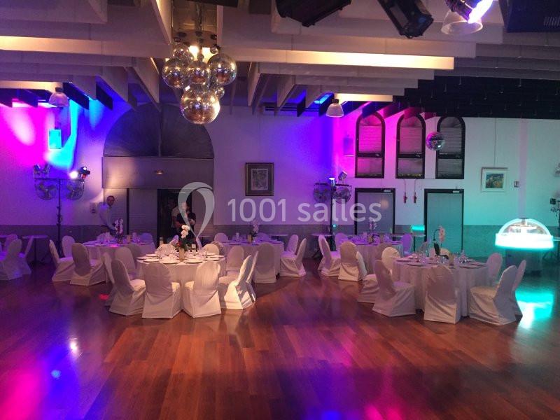 Salle de réception avec tables rondes dressées, chaises blanches et éclairage coloré.