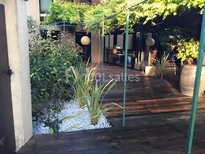 Passage en bois entouré de plantes, menant à une entrée vitrée sous une pergola couverte de végétation.