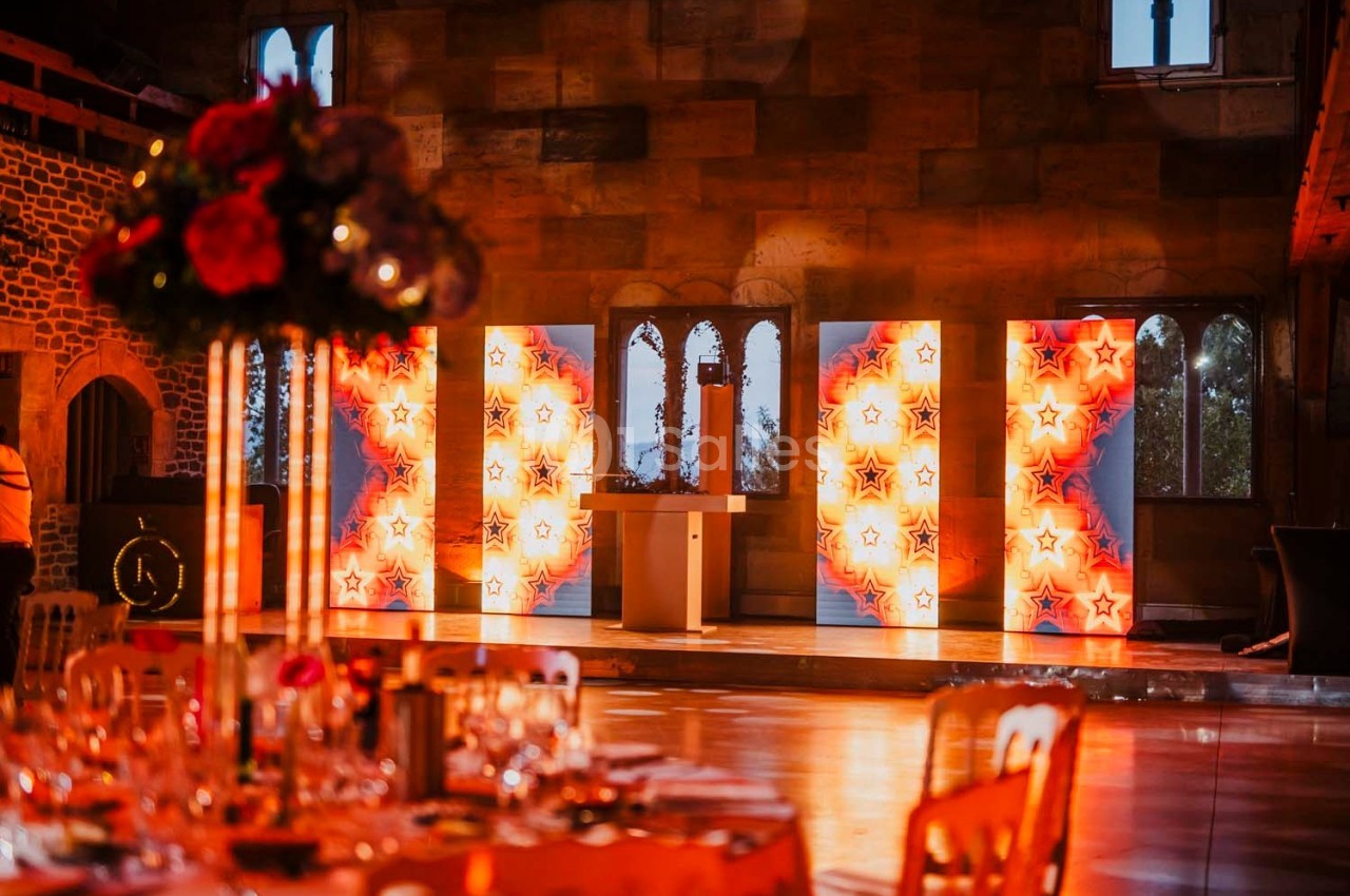 Salle de réception décorée avec des tables dressées et des panneaux lumineux étoilés devant des fenêtres gothiques.