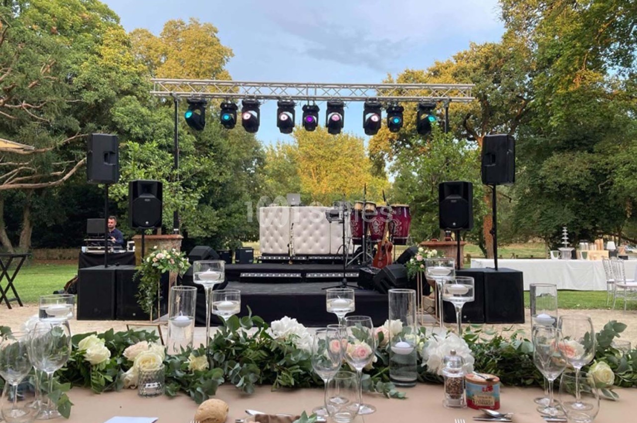 Scène extérieure avec éclairage, instruments de musique et table décorée au premier plan, entourée de verdure.