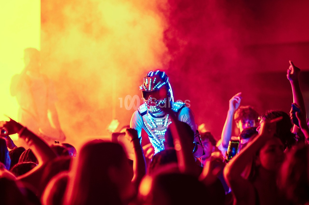 Un artiste en costume lumineux se produit sur scène entouré d'une foule dans une ambiance colorée et enfumée.