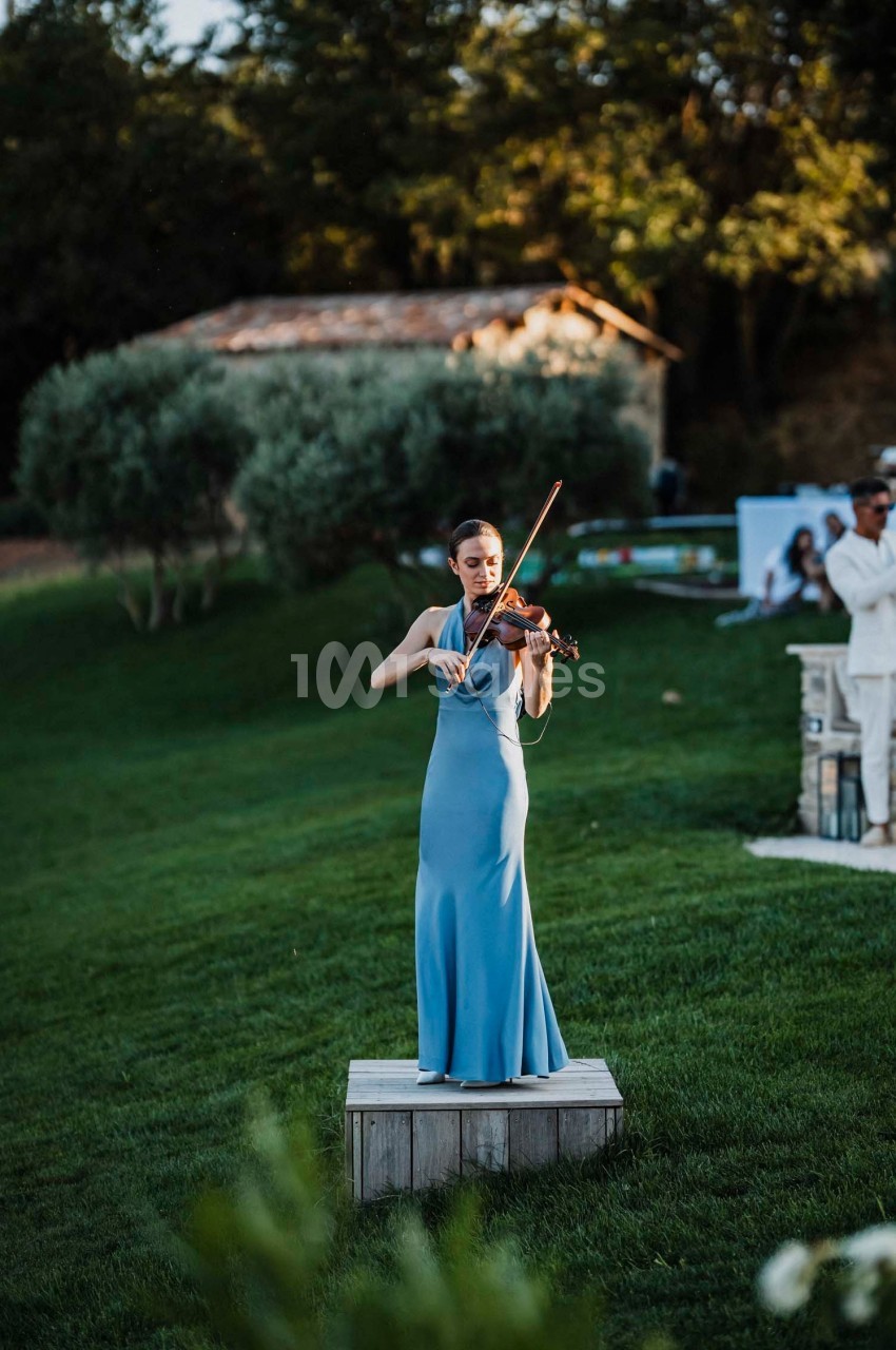 Une femme en robe bleue joue du violon sur une petite estrade en bois dans un jardin verdoyant.