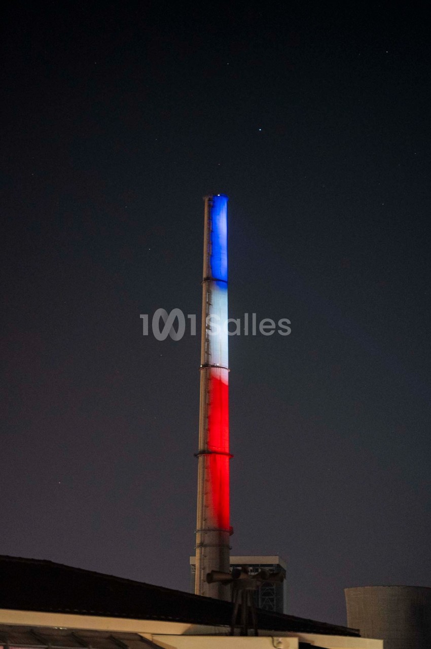 Cheminée industrielle illuminée en bleu, blanc et rouge dans un paysage nocturne.
