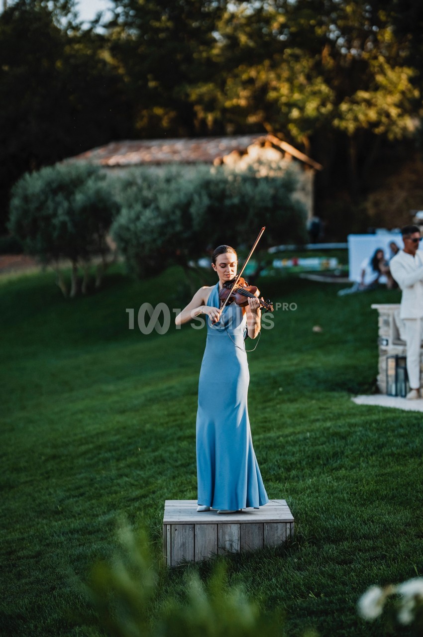 Une femme en robe bleue joue du violon sur une petite estrade en bois dans un jardin verdoyant.