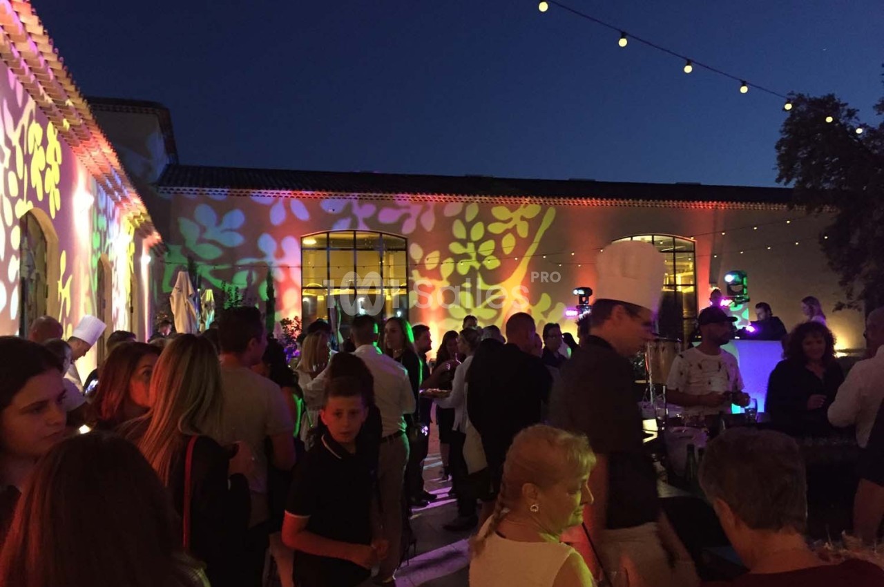 Groupe de personnes rassemblées en soirée festive en extérieur, éclairée par des lumières colorées projetées sur un bâtiment.
