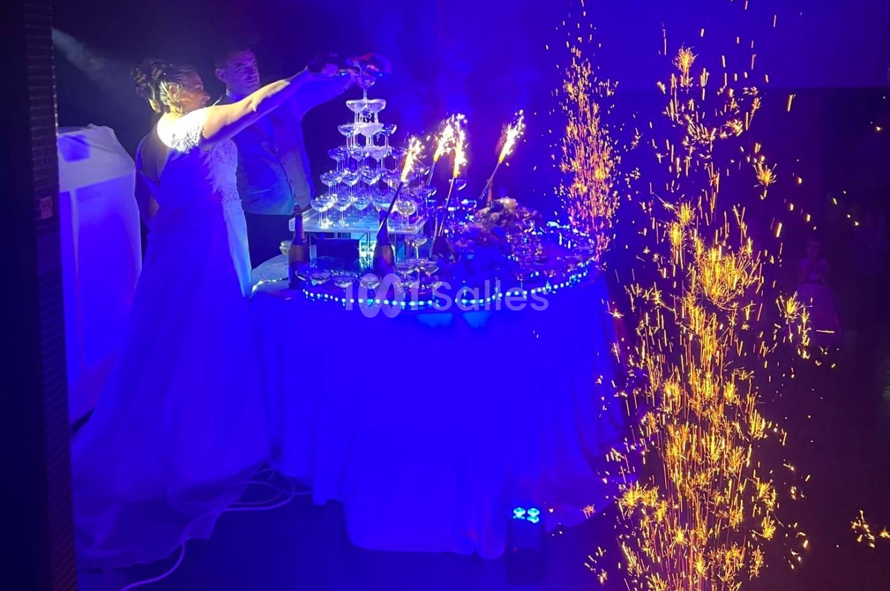 Une mariée verse du champagne dans une fontaine de verres illuminée, entourée de décorations et de gerbes d'étincelles.