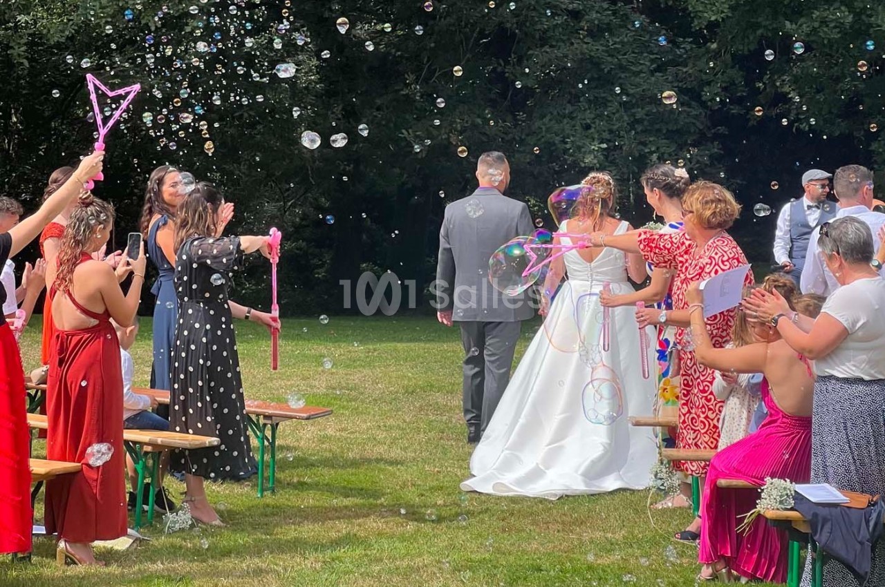 Des invités soufflent des bulles de savon autour d'un couple en tenue de mariage avançant sur une pelouse.