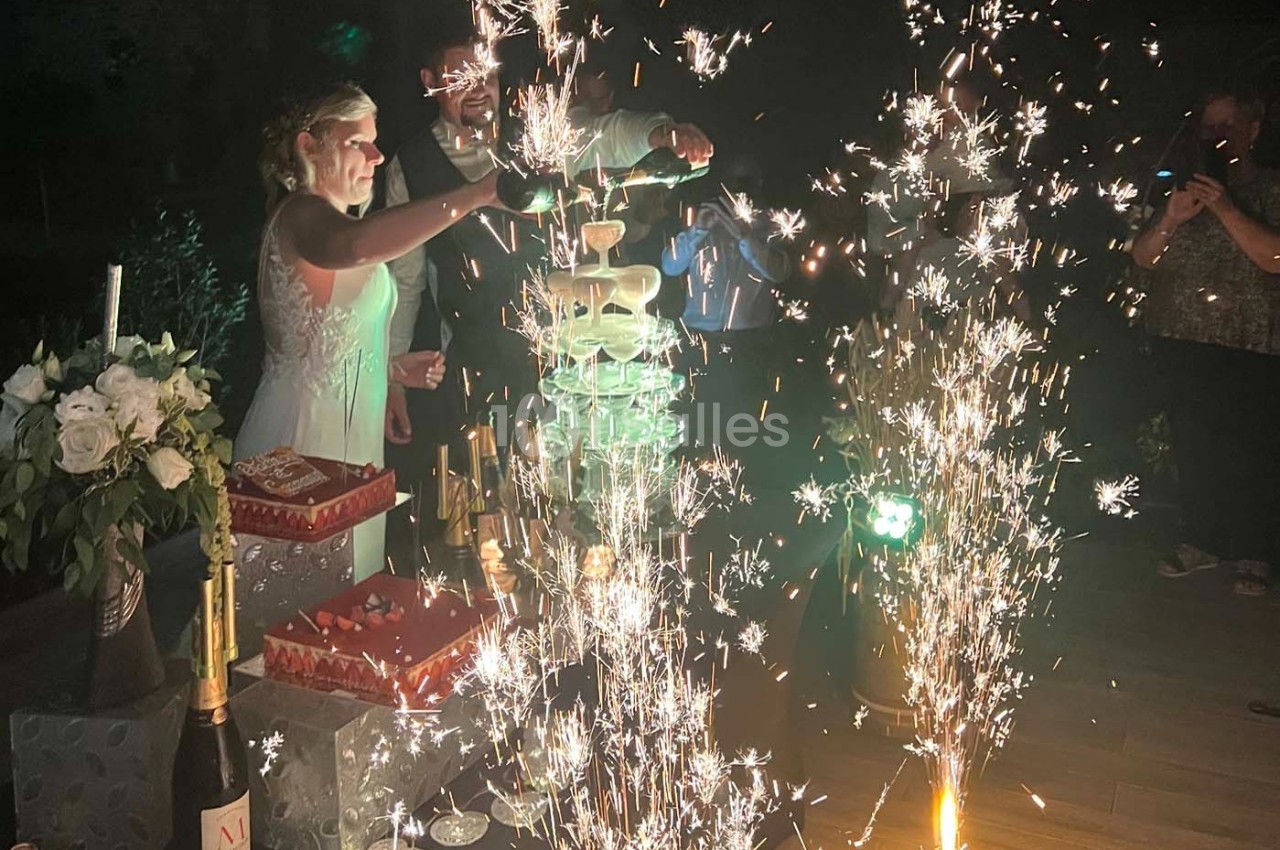 Une mariée verse du champagne dans une pyramide de verres entourée de feux d'artifice et de décorations festives.