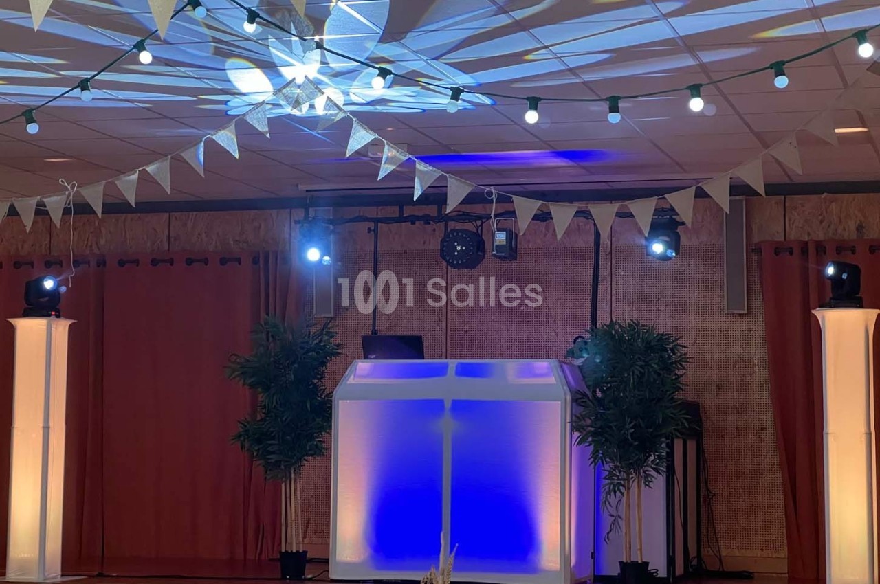Scène décorée avec guirlandes lumineuses, plantes et une table de DJ éclairée en bleu et blanc.