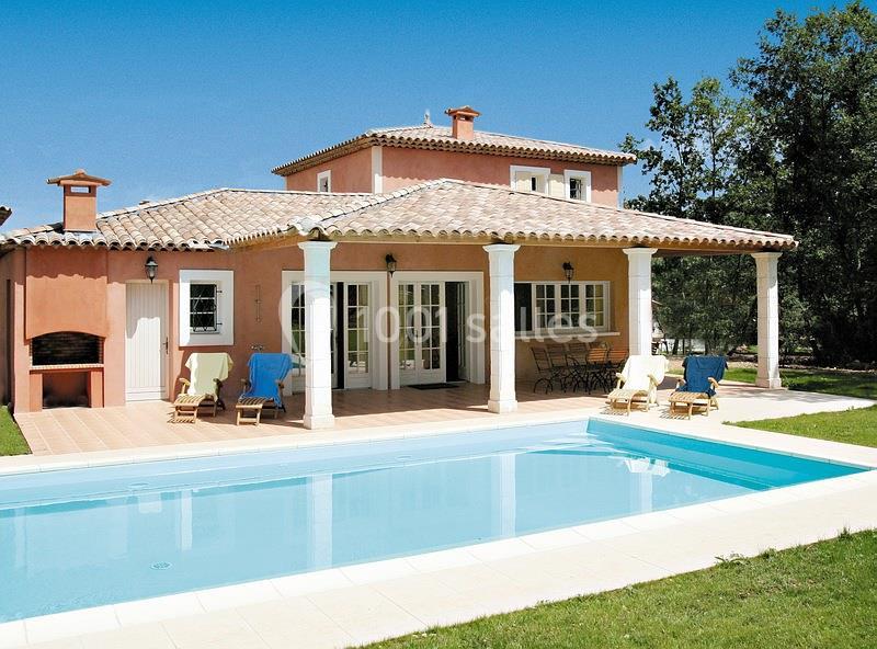 Maison méditerranéenne avec piscine, terrasse, chaises longues et jardin arboré sous un ciel bleu.