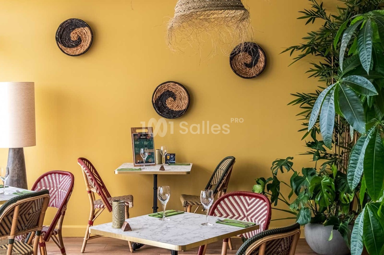 Salle de restaurant avec murs jaunes, tables en marbre, chaises en rotin et plantes vertes en décoration.