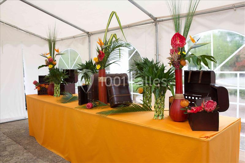 Décoration tropicale avec vases rouges, fleurs exotiques et coffres en bois sur une table recouverte d'une nappe orange.