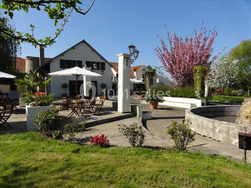 Terrasse extérieure avec tables, chaises, parasols, fontaine et jardin fleuri sous un ciel dégagé.