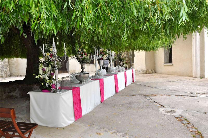 Table décorée avec nappes blanches et roses, ornée de chandeliers et fleurs, placée sous un grand arbre.