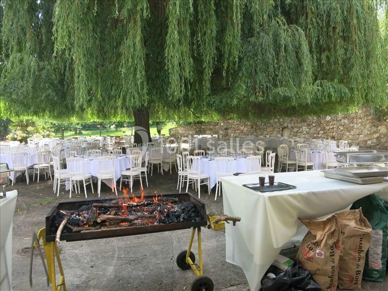Barbecue en plein air avec des tables dressées sous un grand arbre, dans un cadre verdoyant et champêtre.