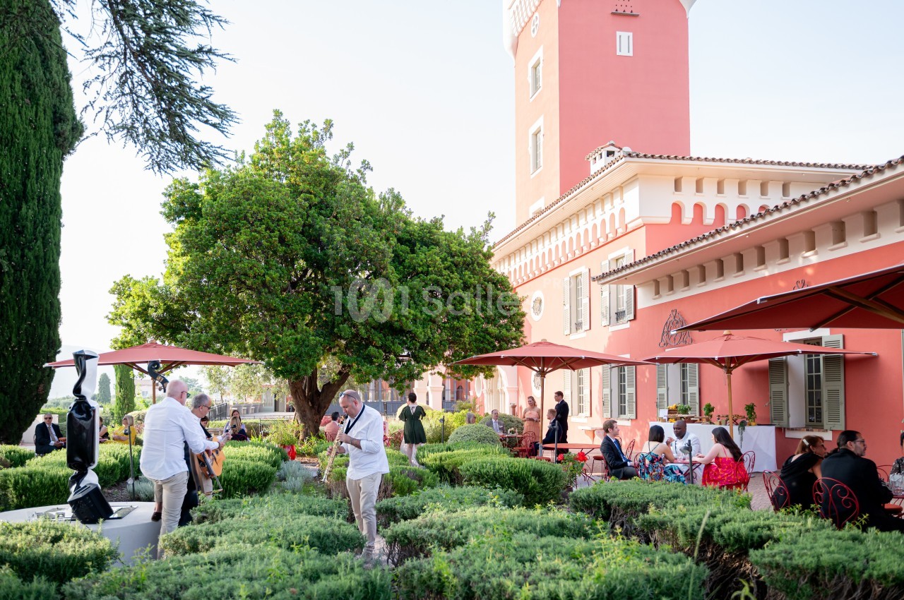 Des personnes profitent d'un jardin avec des tables, des parasols rouges et un bâtiment rose en arrière-plan.