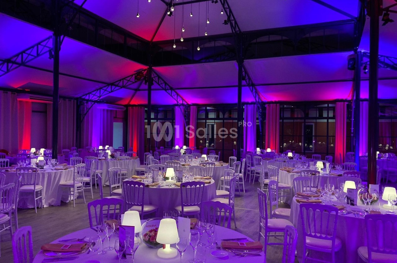 Salle de réception élégamment décorée avec des tables rondes, nappes blanches et éclairage violet et rose.
