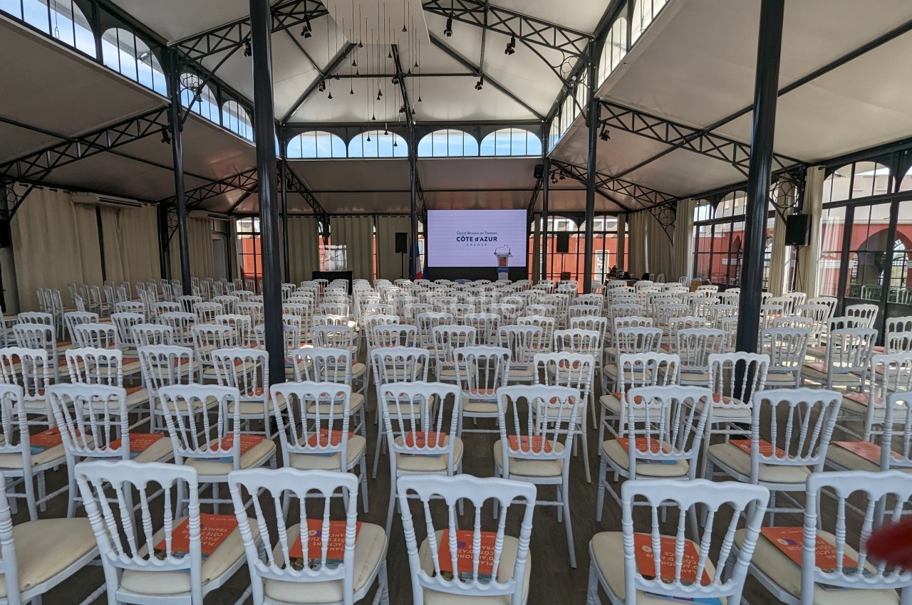 Salle de conférence lumineuse avec des rangées de chaises blanches alignées face à un écran de projection.