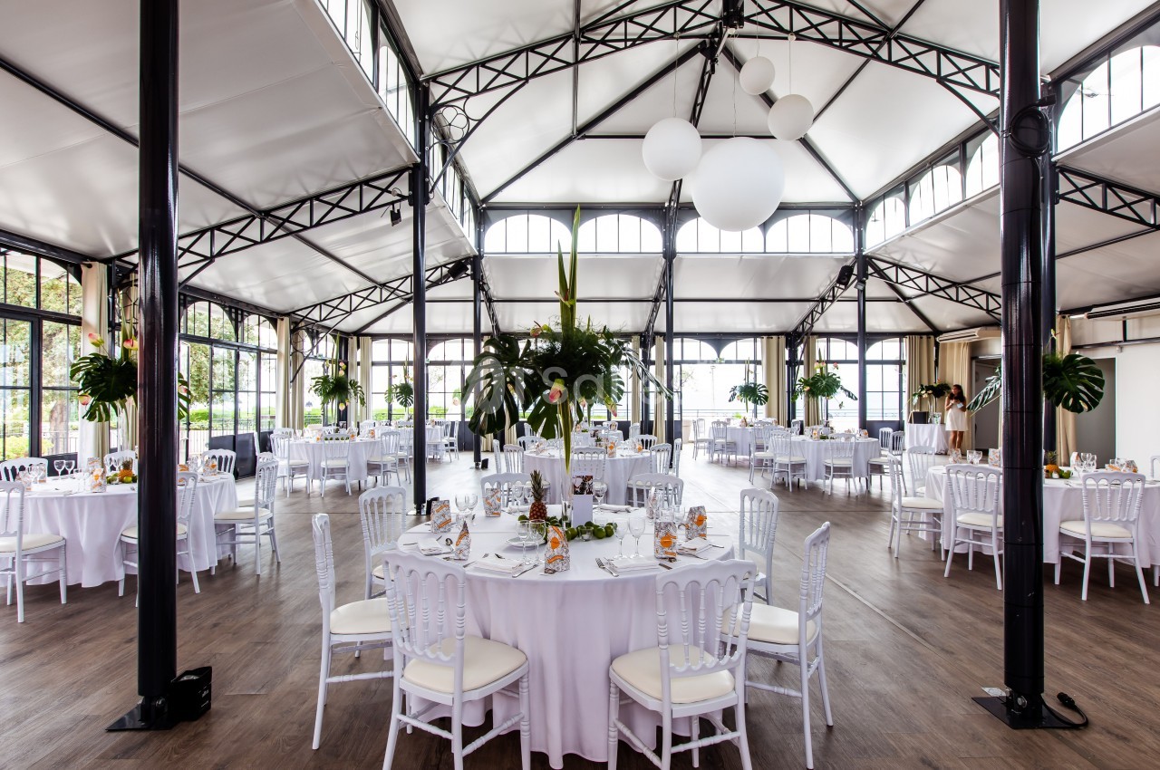 Salle de réception lumineuse avec tables rondes dressées, chaises blanches et décorations végétales.