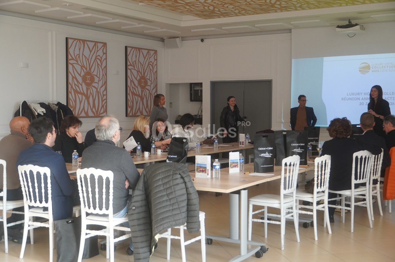 Personnes assises autour de tables lors d'une réunion dans une salle lumineuse avec une présentation projetée.