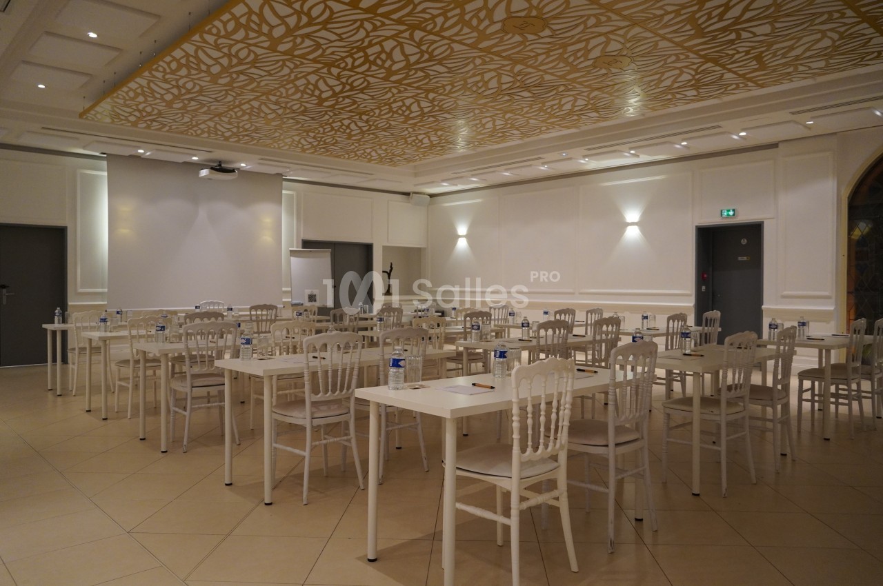 Salle de réunion lumineuse avec des tables blanches alignées, des chaises et un plafond décoré de motifs dorés.