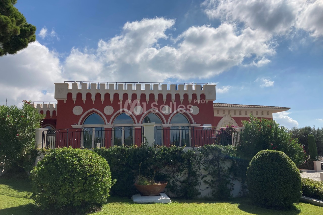 Façade d'un bâtiment rouge avec des arcs et des créneaux, entouré de verdure sous un ciel partiellement nuageux.