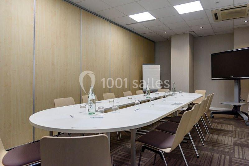 Salle de réunion avec une table ovale, des chaises, un tableau de conférence, une télévision et des bouteilles d'eau.