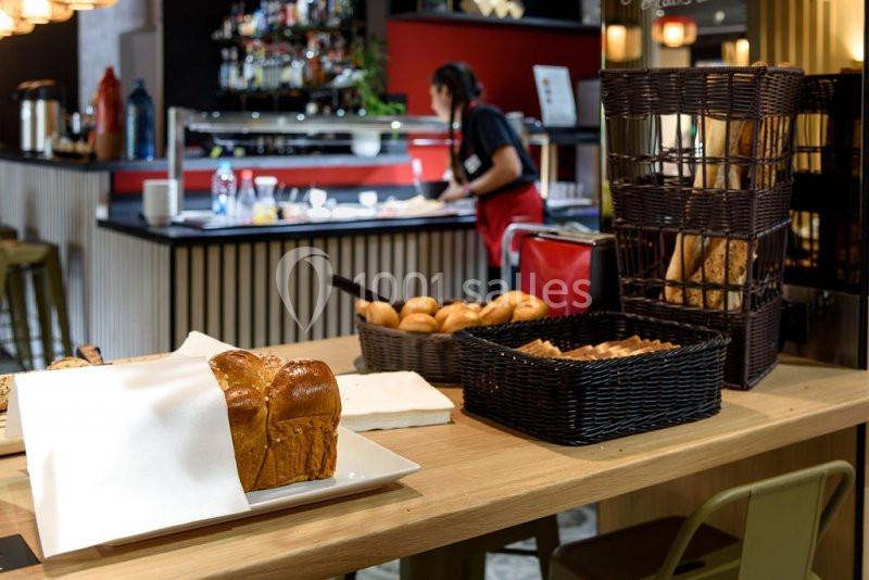 Pain et viennoiseries disposés sur une table en bois, avec une employée en arrière-plan près d'un buffet.
