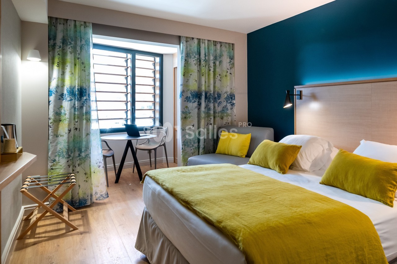 Chambre lumineuse avec lit double, coussins jaunes, bureau près de la fenêtre et rideaux à motifs floraux.