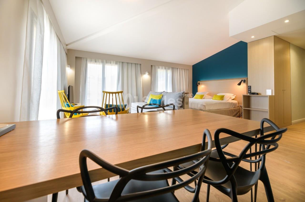Salle lumineuse avec table en bois, chaises modernes, coin salon, lit double et murs aux tons neutres et bleu.