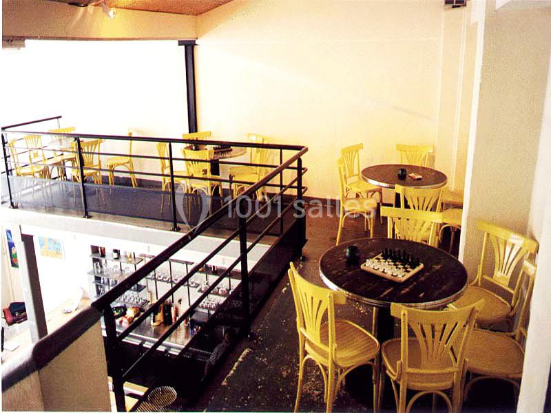 Salle d'un café avec mezzanine, tables rondes, chaises en bois clair et un échiquier posé sur une table.