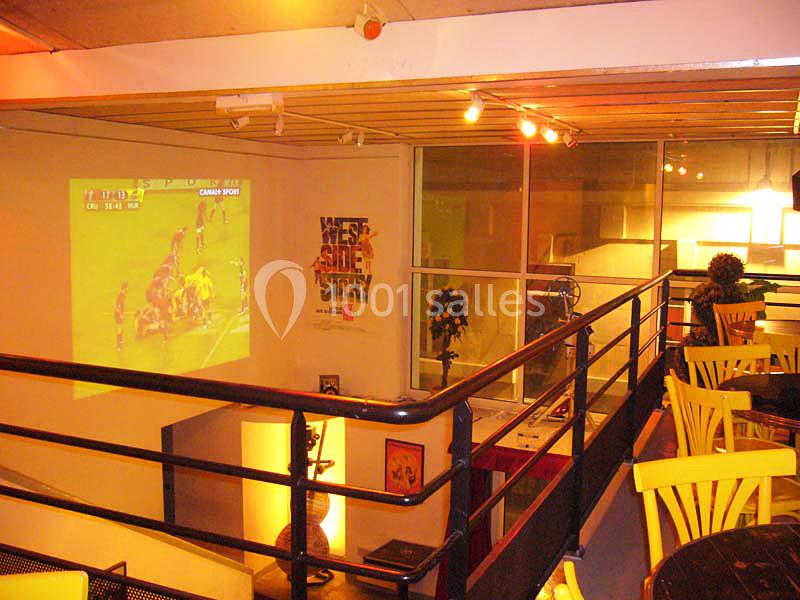 Salle avec mezzanine, tables et chaises jaunes, projection d'un match de rugby sur un mur, éclairage chaleureux.