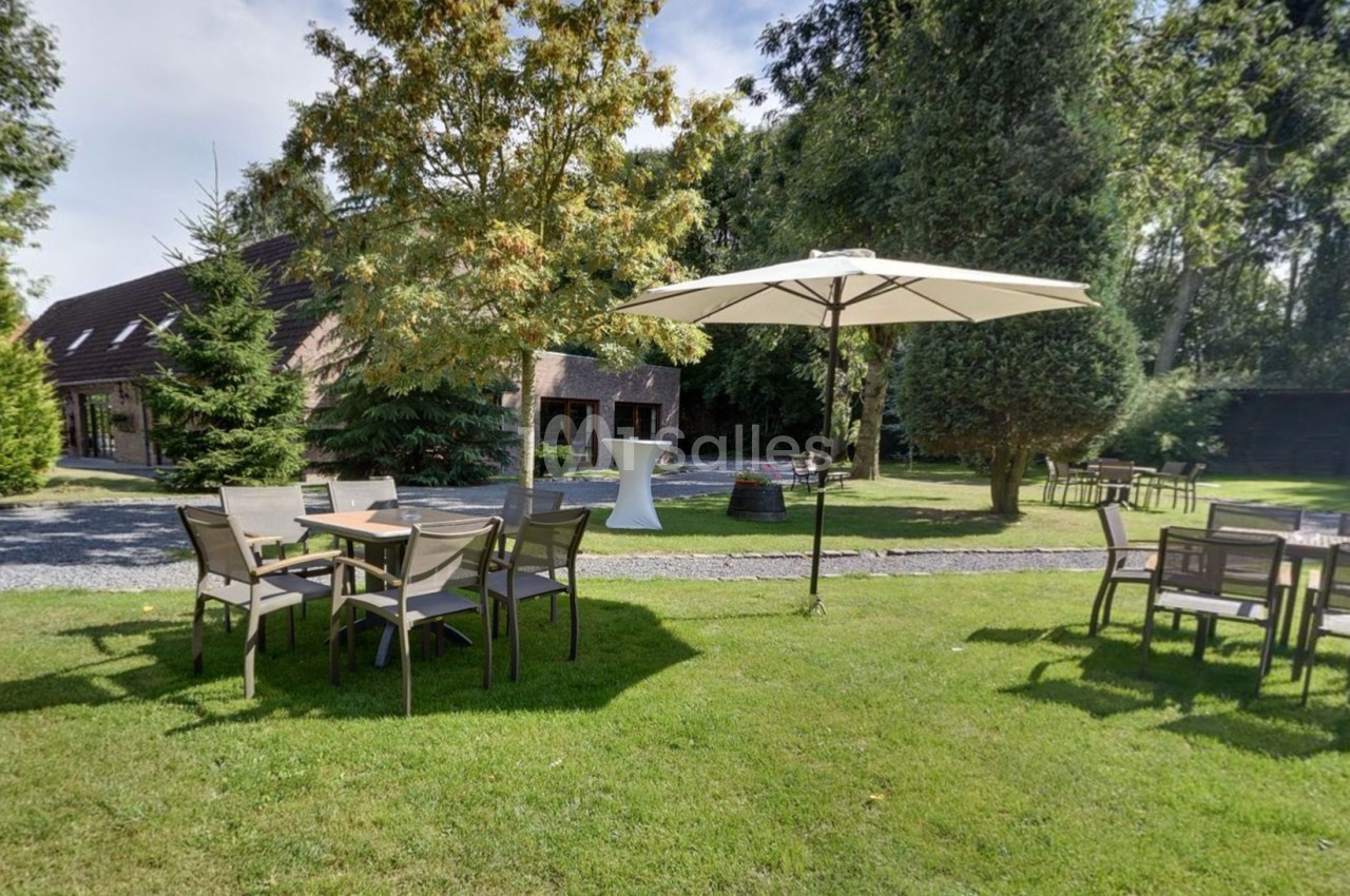 Jardin avec tables, chaises et parasol sur pelouse, entouré d'arbres et d'une maison en arrière-plan.