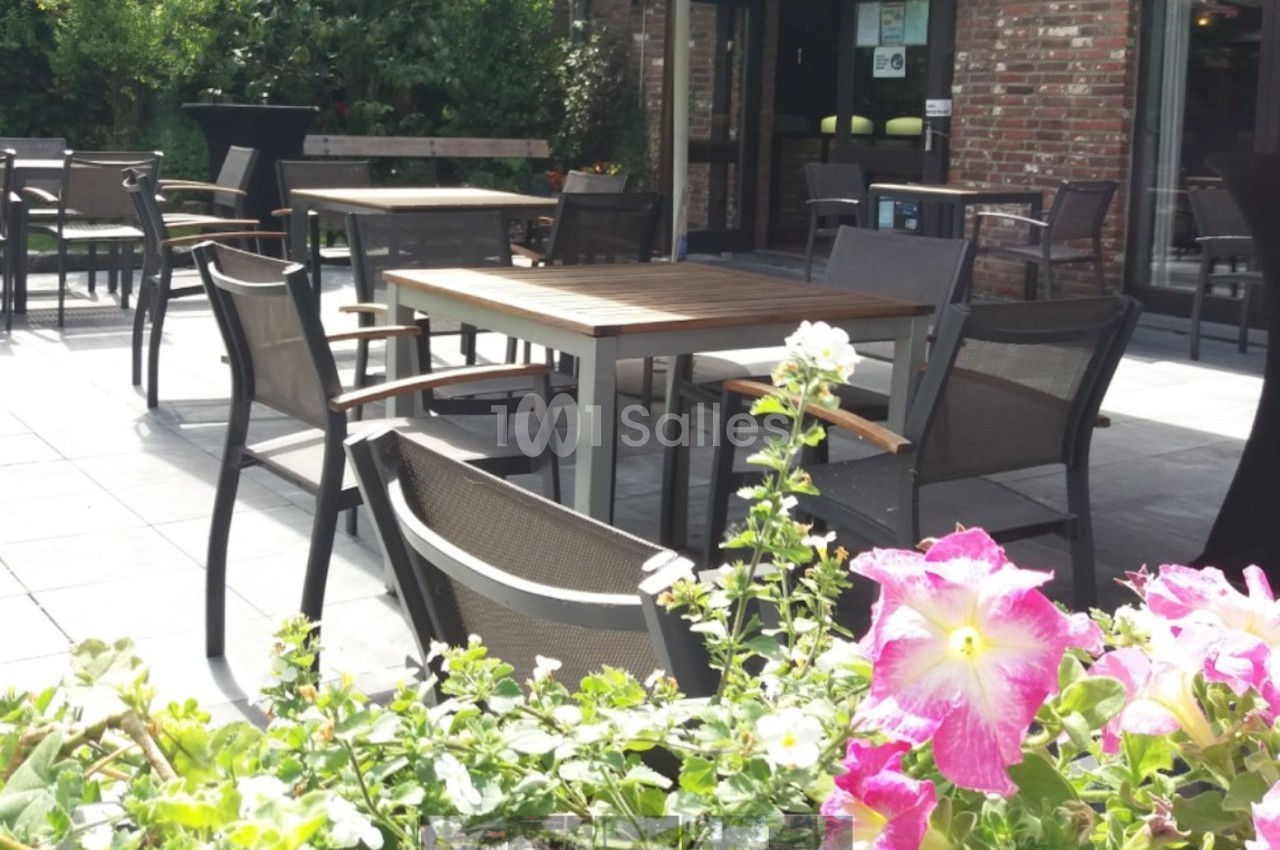 Terrasse extérieure avec tables et chaises en bois et métal, entourée de verdure et de fleurs colorées au premier plan.