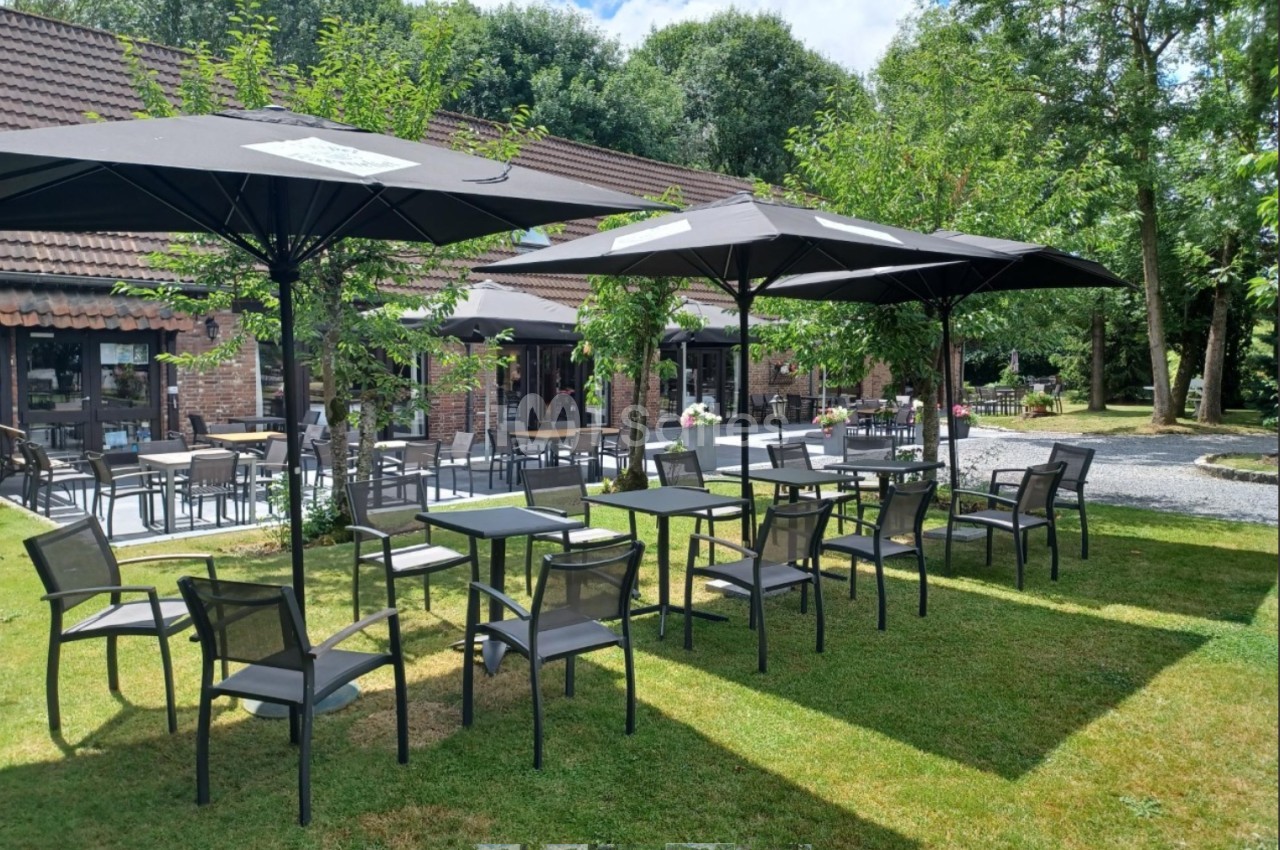 Terrasse extérieure avec tables, chaises et parasols noirs, entourée de verdure et bâtiments en briques.