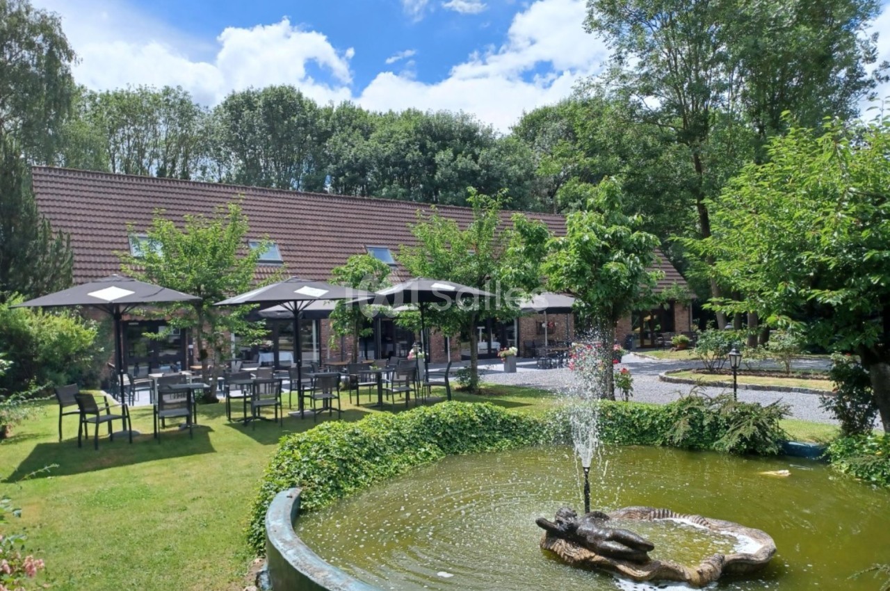 Terrasse extérieure avec tables et parasols, entourée de verdure, près d'un bassin avec une fontaine.