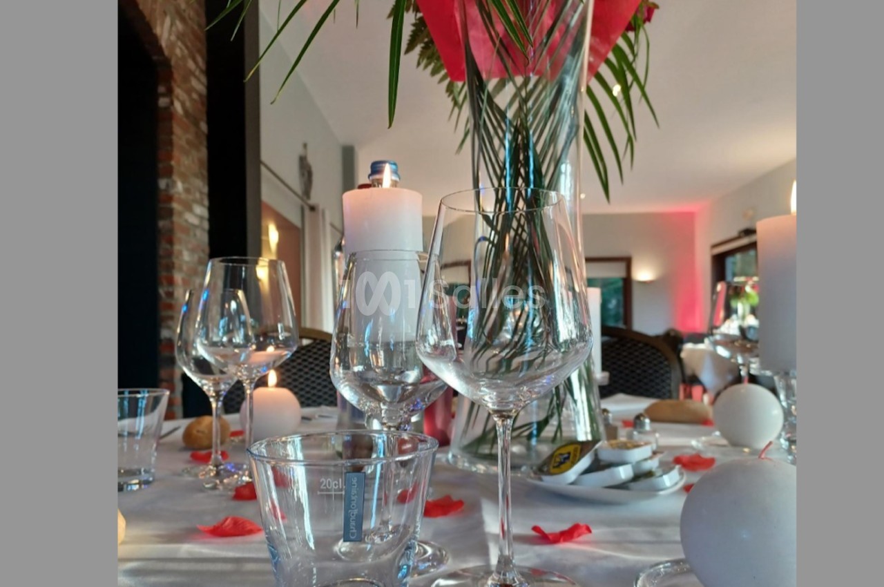 Table dressée avec des verres, bougies et pétales rouges, décorée d'un grand vase avec des feuilles et fleurs.