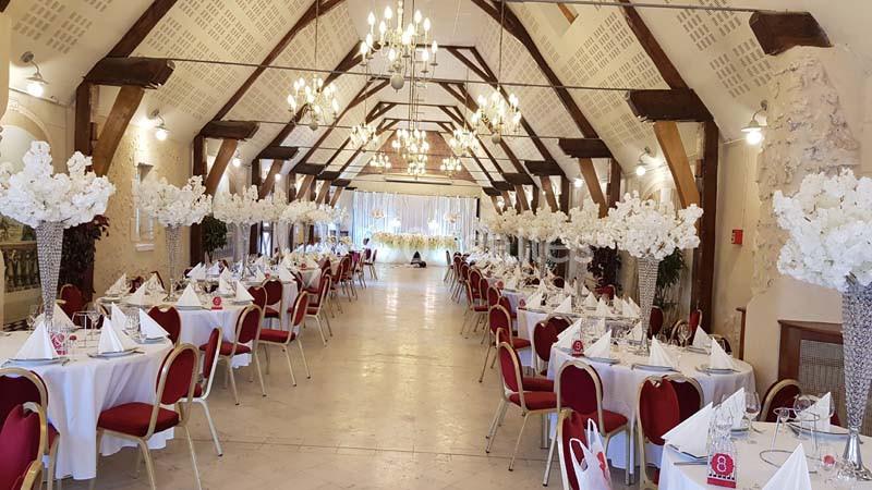 Salle de réception élégante avec tables dressées, chaises rouges et décorations florales blanches sous des lustres suspendus.