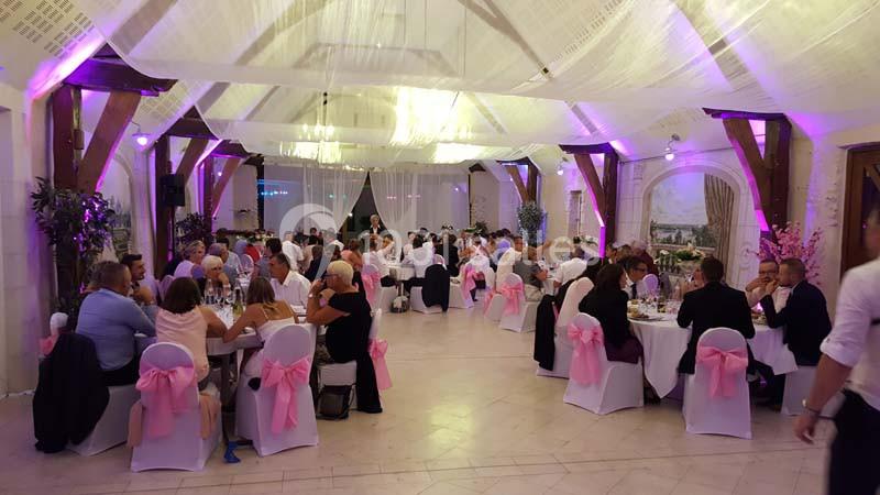 Salle de réception décorée avec des nappes blanches et des rubans roses, accueillant des invités assis à des tables rondes.