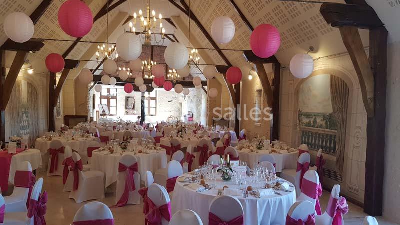 Salle de réception décorée avec des tables rondes, nappes blanches, chaises ornées de nœuds roses et lanternes suspendues.