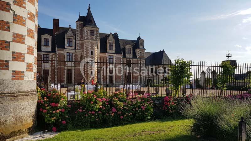 Façade d'un château en pierre avec des toits en ardoise, entouré de fleurs et d'une pelouse sous un ciel dégagé.