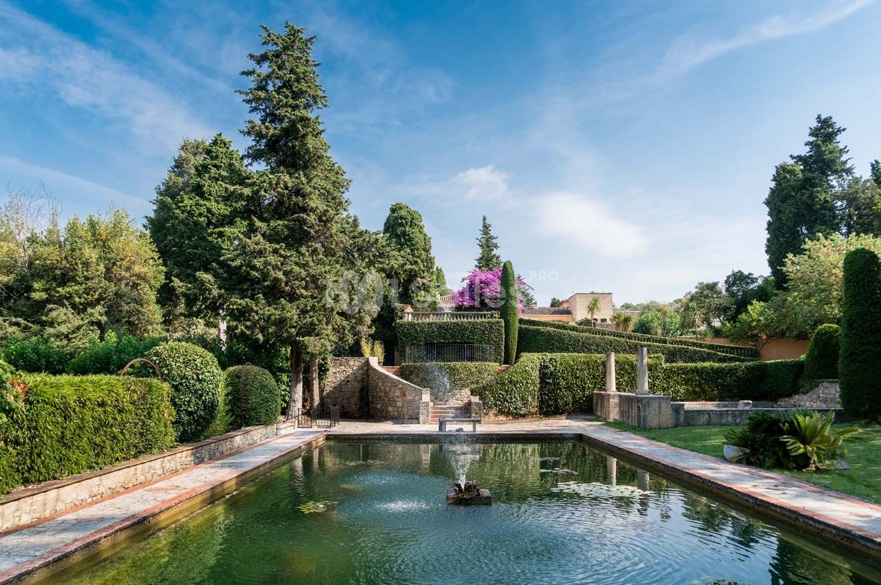 Jardin avec bassin central, fontaine, arbres et buissons taillés, sous un ciel dégagé.