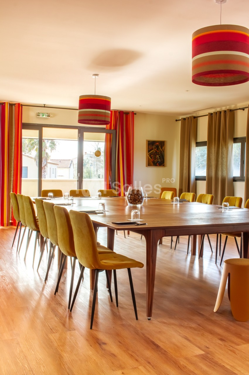 Salle lumineuse avec une grande table en bois entourée de chaises jaunes, rideaux colorés et suspensions rayées.