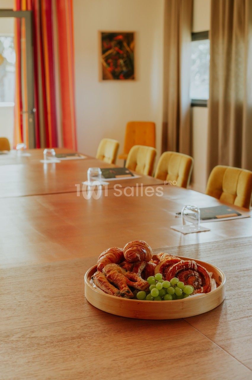 Plateau de viennoiseries et raisins posé sur une grande table en bois entourée de chaises jaunes.