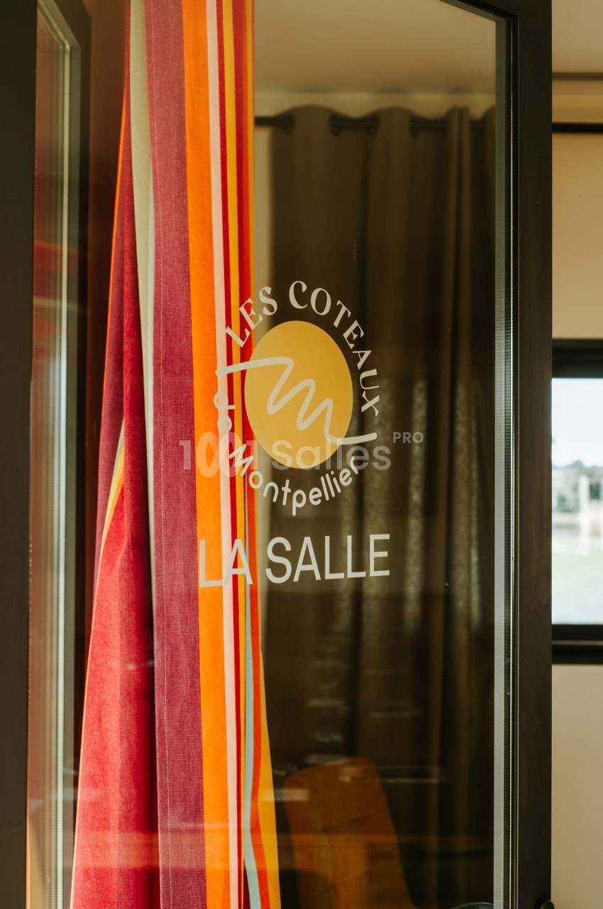 Porte vitrée avec un logo circulaire ’Les Coteaux Montpellier’ et des rideaux colorés en arrière-plan.