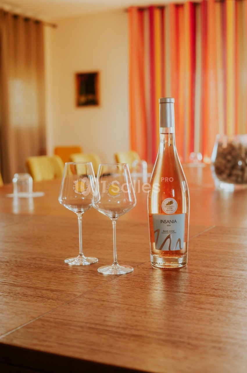Bouteille de vin rosé et deux verres sur une table en bois, avec un fond de rideaux colorés et des chaises jaunes.