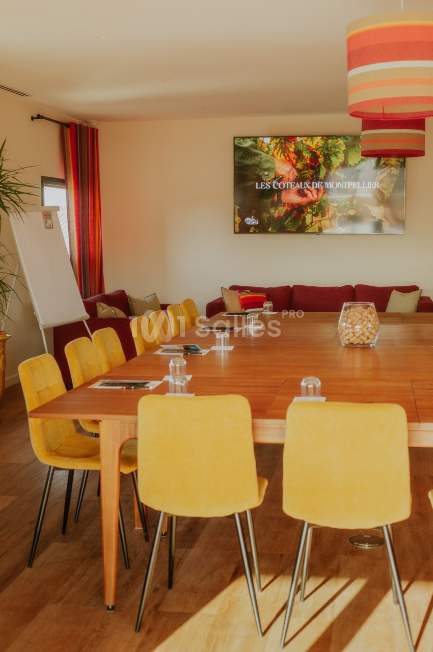 Salle de réunion lumineuse avec table en bois, chaises jaunes, écran affichant ’Les Coteaux de Montpellier’ et tableau blanc.