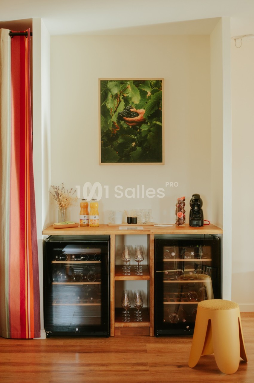 Deux caves à vin, des boissons et une machine à café sur un meuble en bois, avec un tabouret et un tableau au mur.
