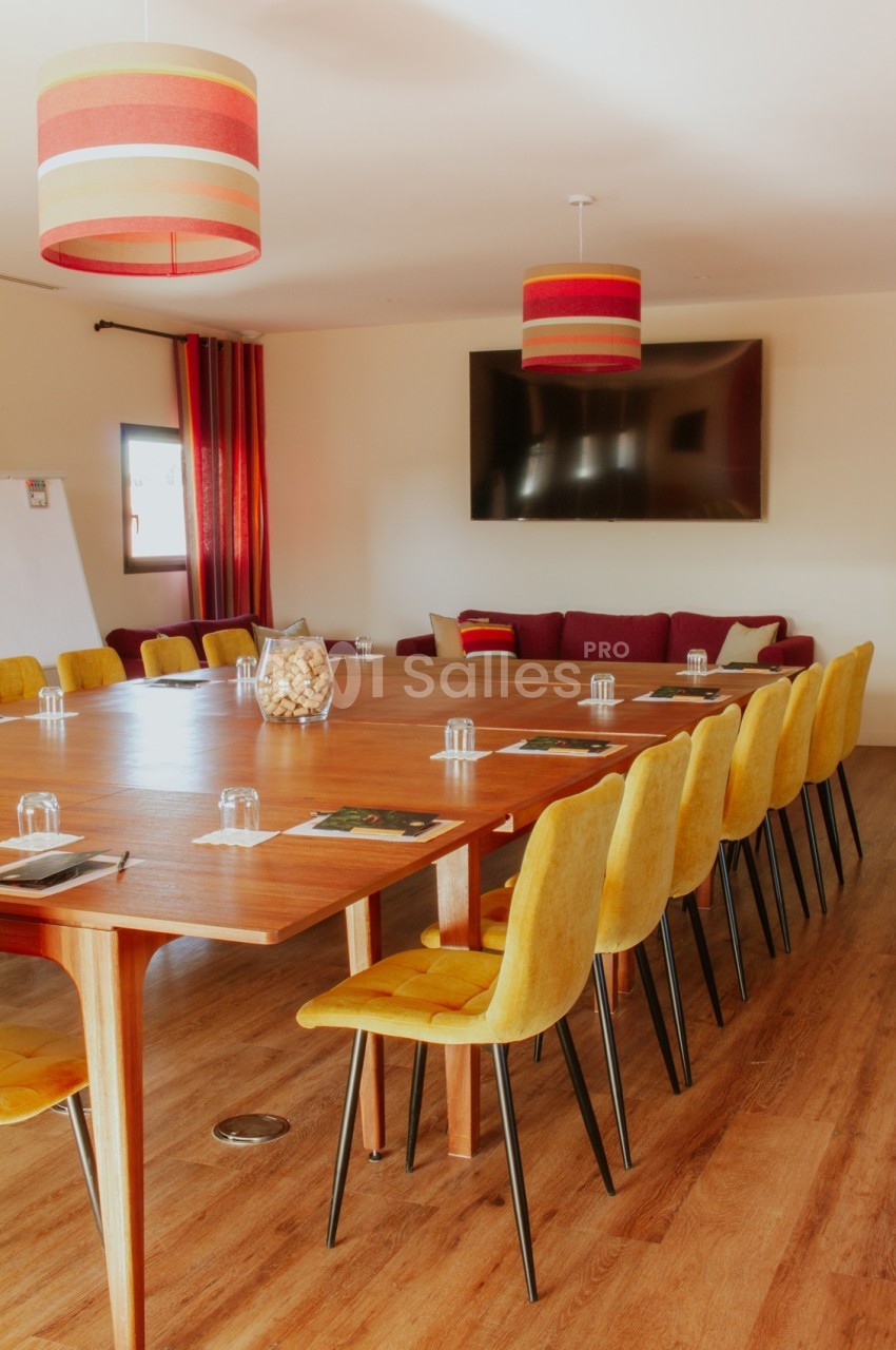 Salle de réunion avec une grande table en bois, des chaises jaunes, un écran mural et des lampes suspendues colorées.